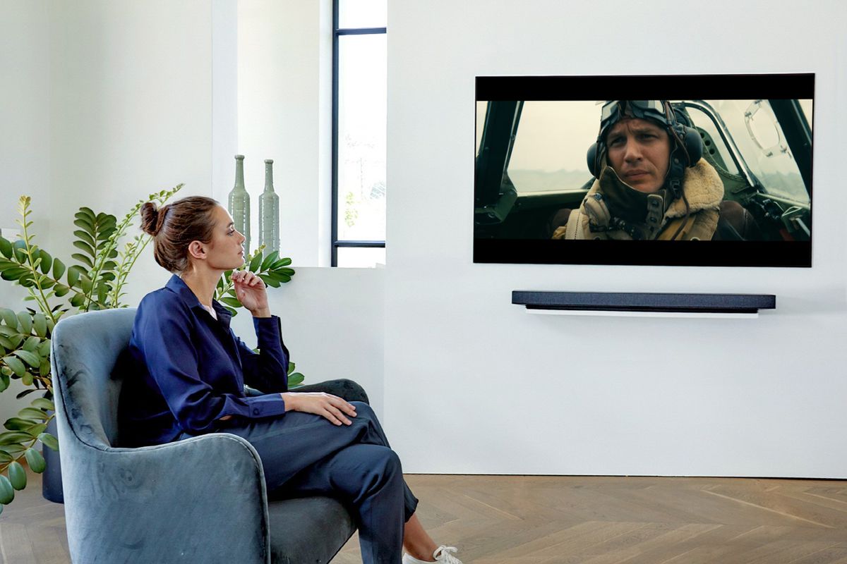 a white woman in a blue shirt and blue pants sits in a blue armchair watching Dunkirk on a wall-mounted LG 4K TV with a wall-mounted soundbar beneath it
