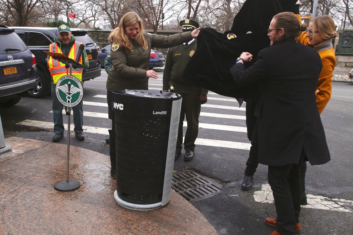 Several individuals stand around a black and grey trash can in a street corner.