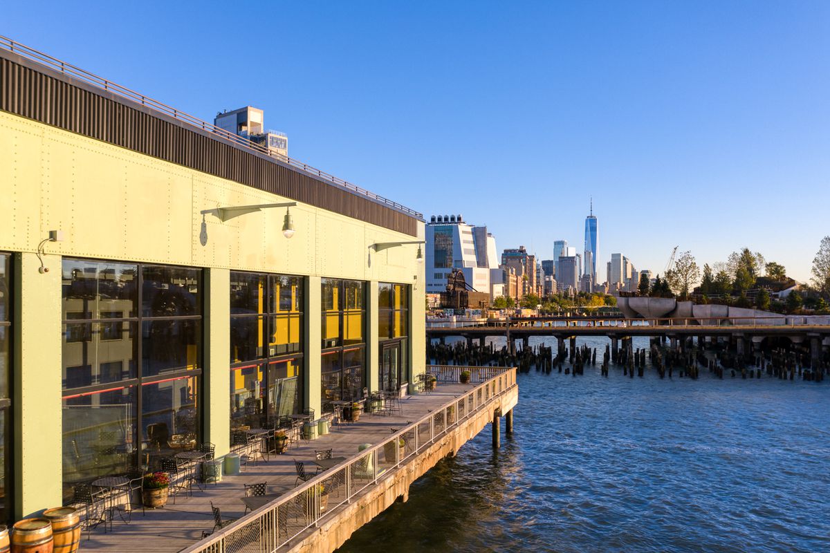 A building on a pier with outdoor dining set up and an NYC building skyline in the background