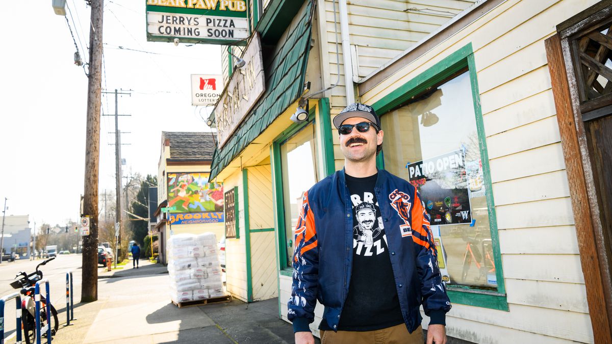 Jerry Benedetto of Jerry’s Pizza — a man with a black handlebar moustache, a Chicago Bears letterman’s jacket, and a baseball cap — stands outside the Bear Paw Inn.