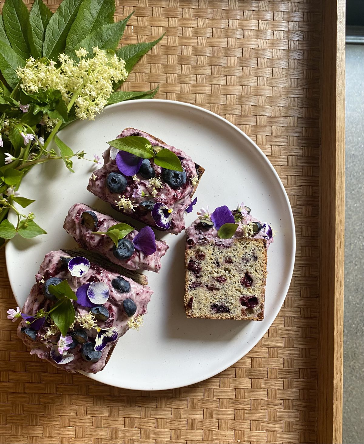 From above, a loaf cake topped with purple icing, whole blueberries, and sprigs of herbs.