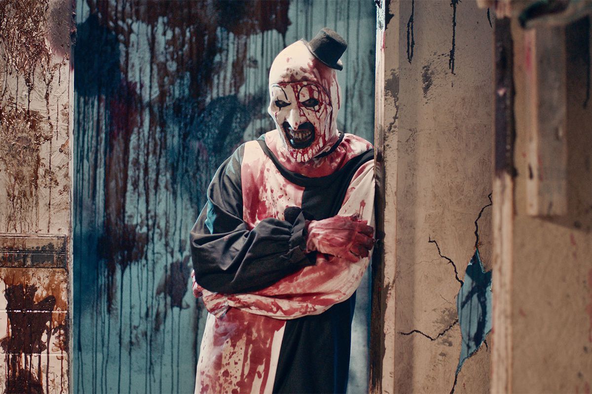 A man in a black and white clown outfit and makeup leaning against a doorframe, splattered with blood and smiling.