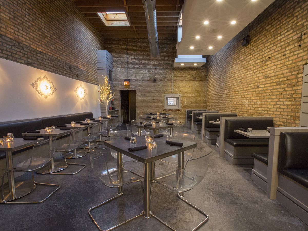 A narrow dining room with exposed brick walls and grey tables and chairs.