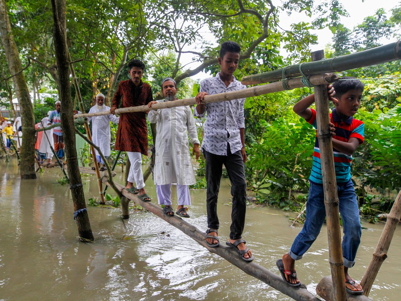 People walking on a makeshift bridge over muddy floodwaters in a wooded area.