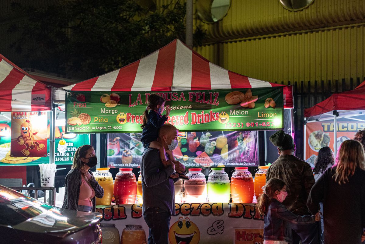 A masked family passes in front of a lit food stand selling drinks.