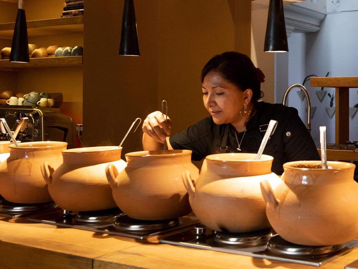 A chef leans over and stirs a clay pot, one in a row of pots lined up on a counter beneath warm pendant lights