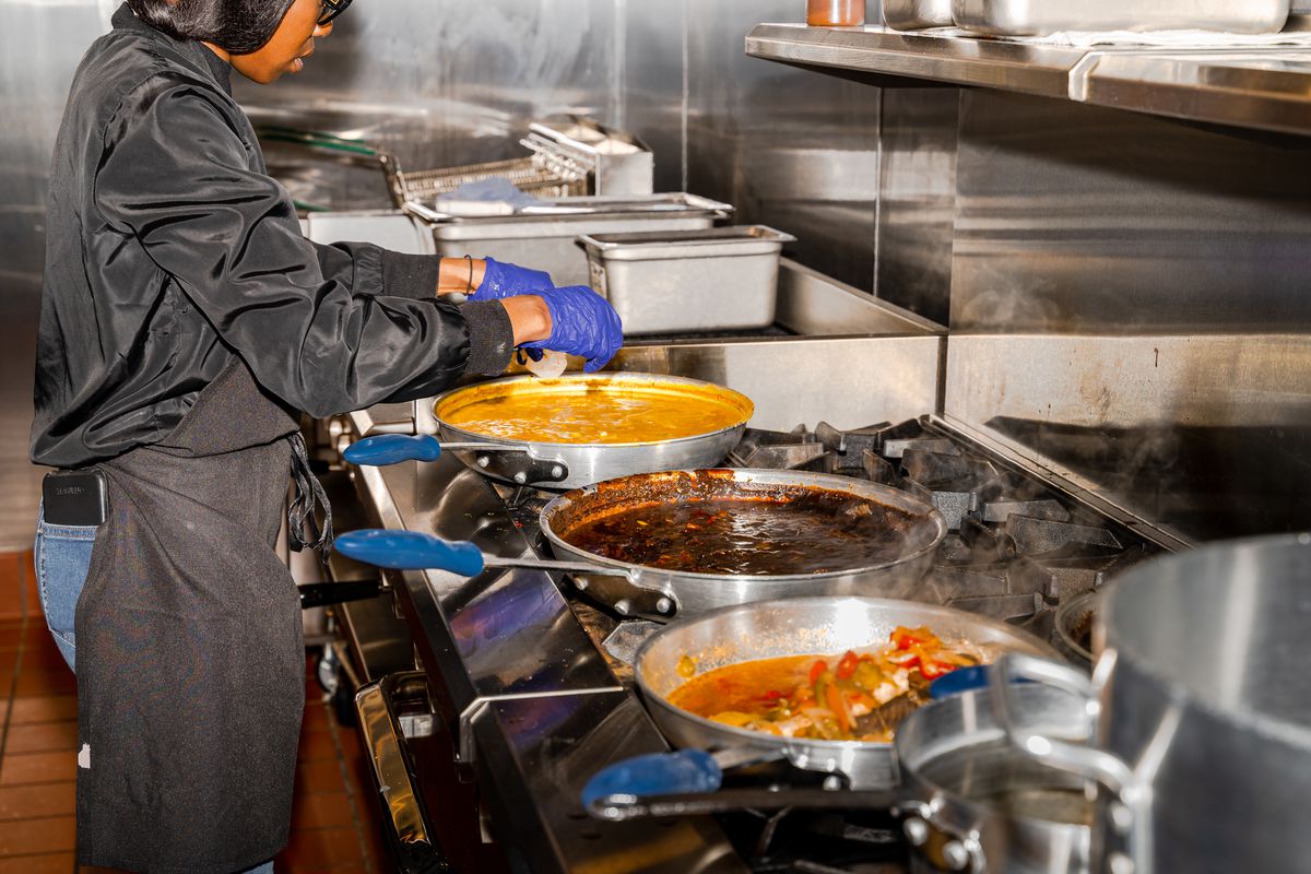 A cook stands over a gas range with three sauté pans filled with food.