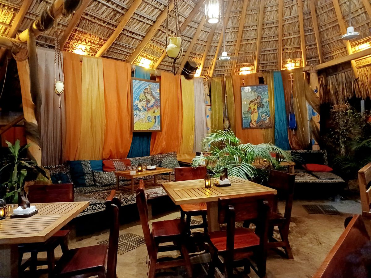 A thatched-roof tent-like space, with pots hung from ropes from the ceiling, Middle Eastern art on the walls, banquettes covered in patterned cushions, and wooden tables surrounded by greenery