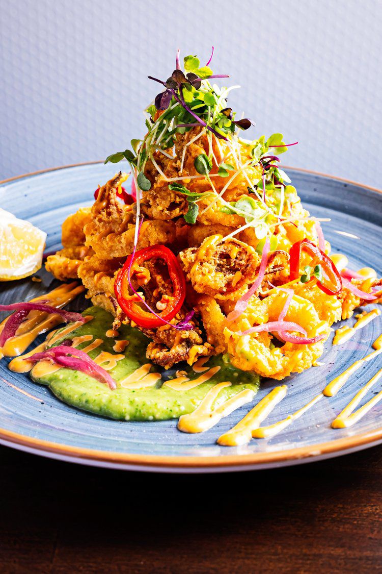 A tower of fried calamari topped with pickled red onion and shoots sits on bed of aji amarillo aioli and serrano sauce.