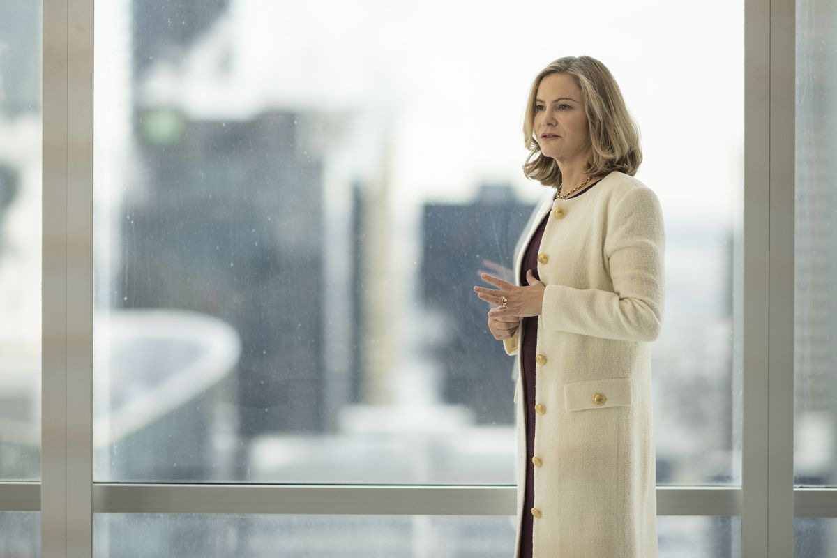 Jennifer Jason Leigh as Lorraine Lyon stands in an expensive white coat in front of an office window in FX’s Fargo season 5.