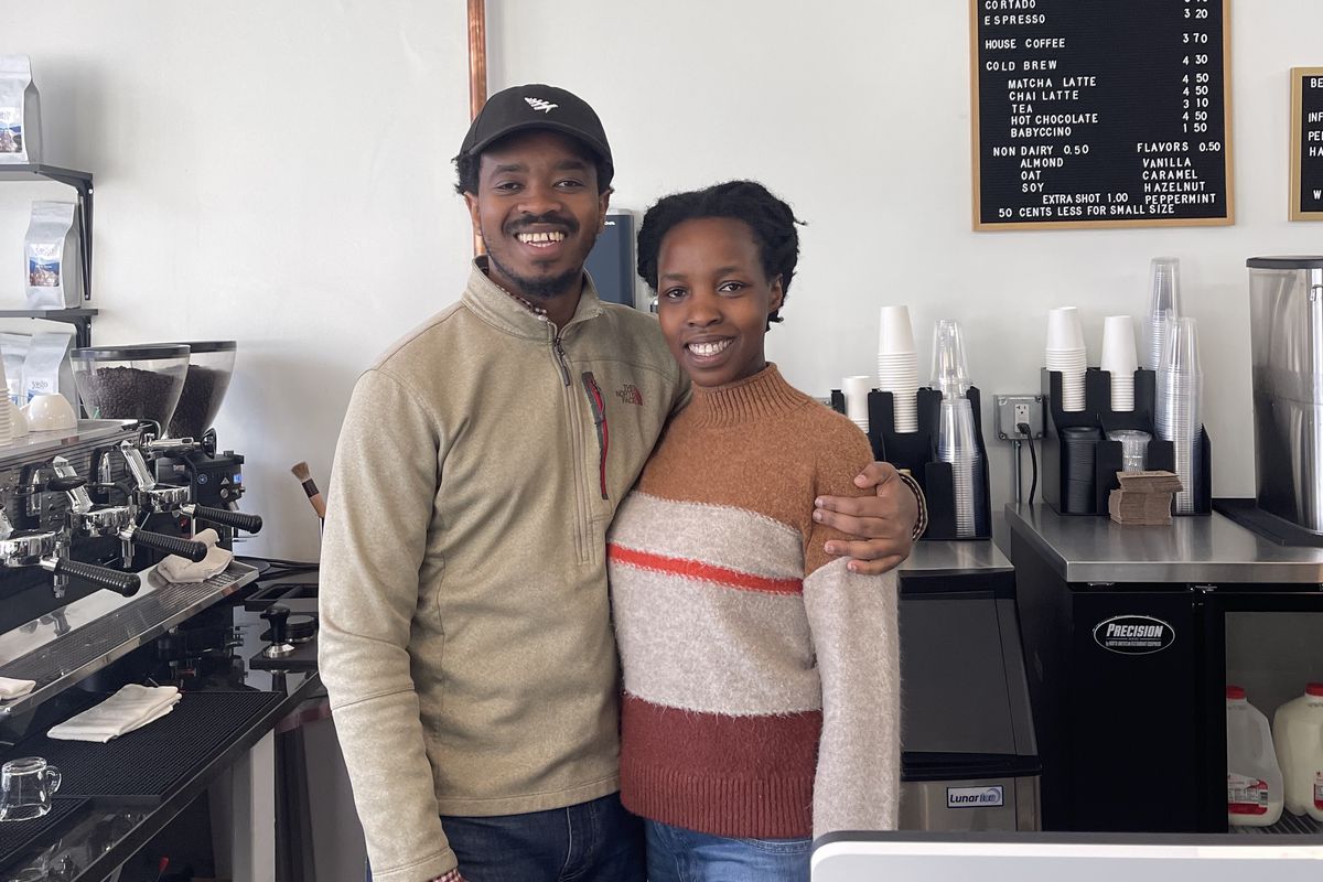 A man and women stand smiling at the camera from behind a coffee bar, with their arms around each other.