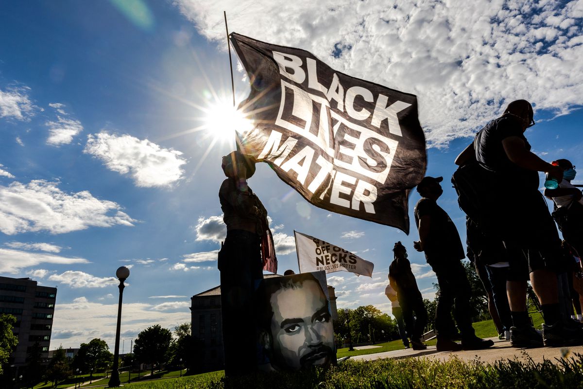 A Black woman holds up a giant black flag, with “Black Lives Matter” written on it in white. She, and the flag, are framed by the sun, which is shining brightly in a blue sky.