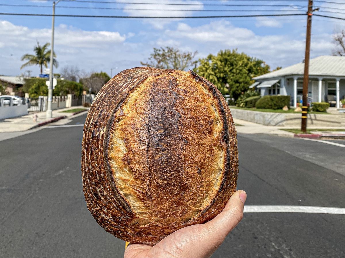 A hand holds up a loaf of bread in the middle of a street during a lightly cloudy day.