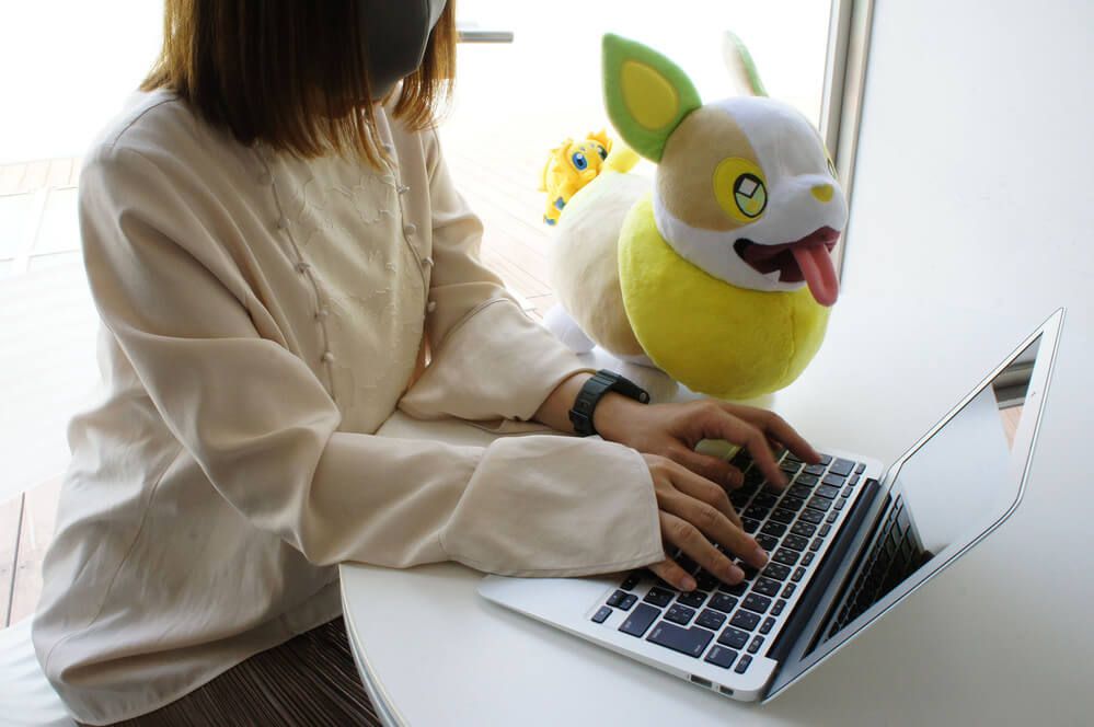 A woman works on a laptop next to a plush toy of Yamper with Joltik attached to its butt