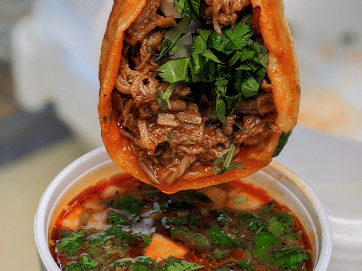 A taco with meat and green leaves being dipped into a broth. 