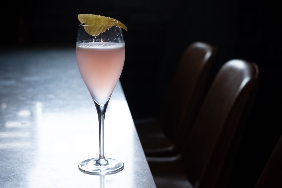 A champagne glass holds a pale pink drink, garnished with lemon on the bar with a beam of light from a nearby window shining on it