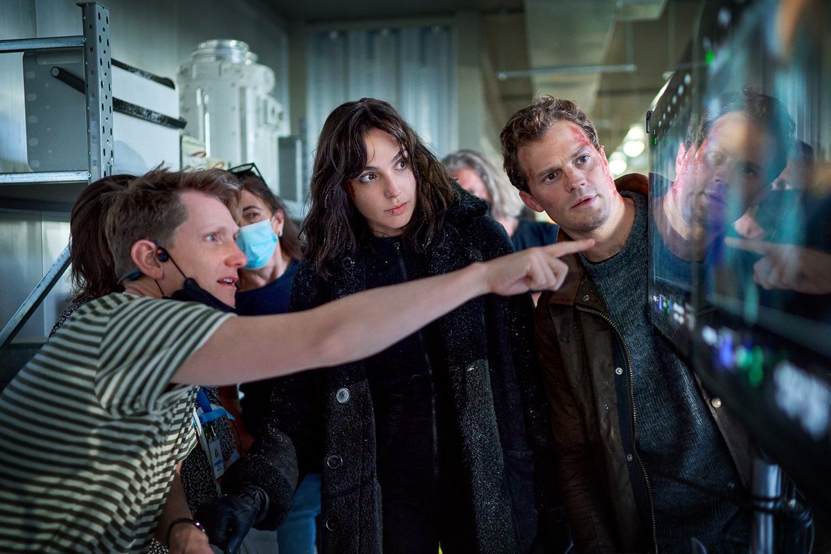 Tom Harper points at a monitor while Gal Gadot, Jamie Dornan, and Jo McLaren (wearing a mask) look on while filming Heart of Stone.