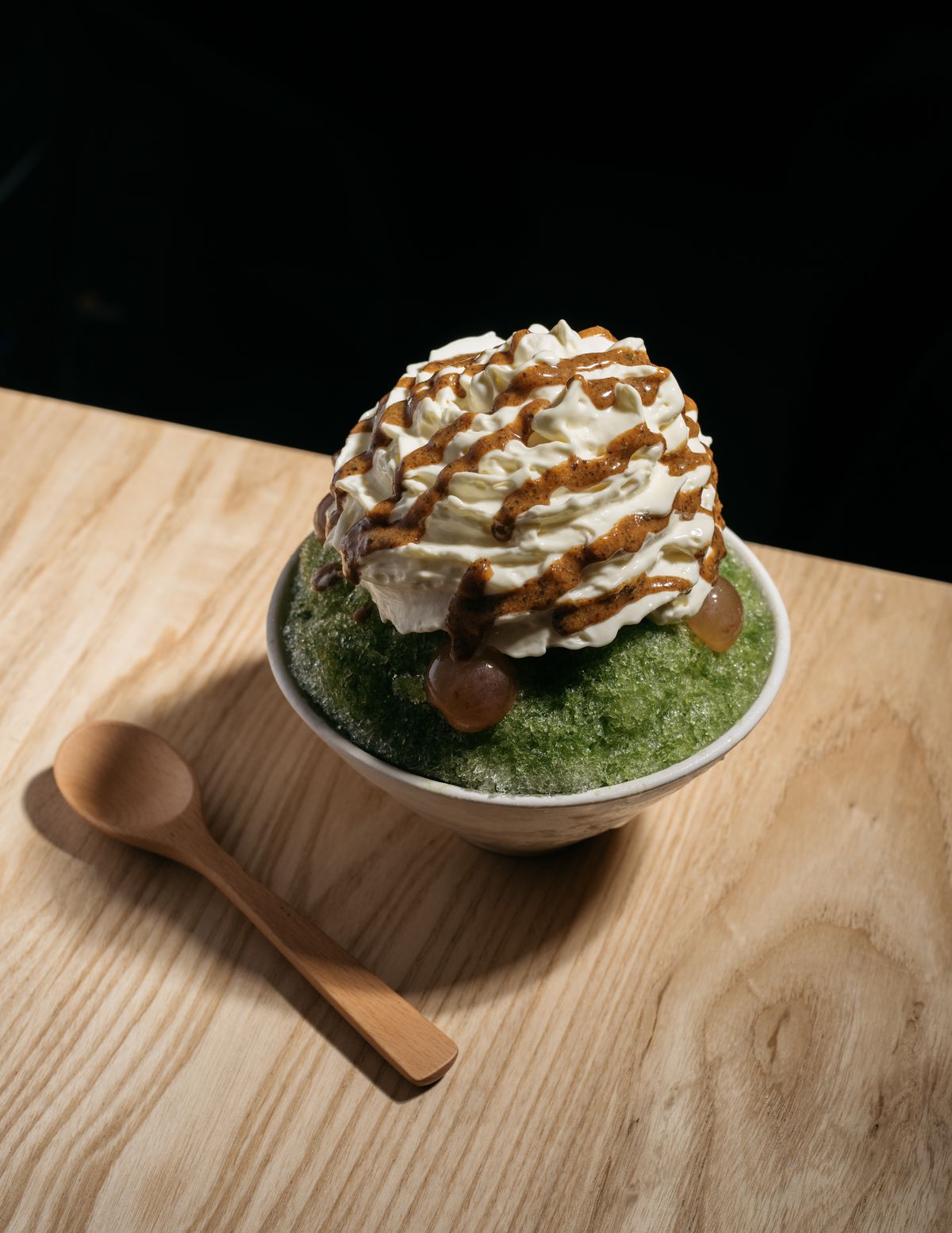 A ceramic bowl full of green shaved ice and topped with cream and drizzled with a caramel sauce. 