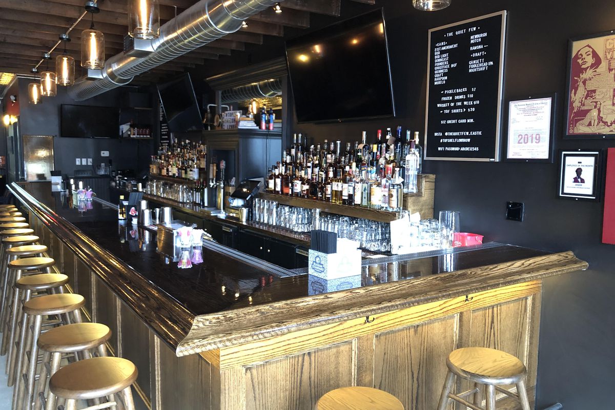 An interior photo of a whiskey bar shows a dark wall and a light wooden bar with stools along it.