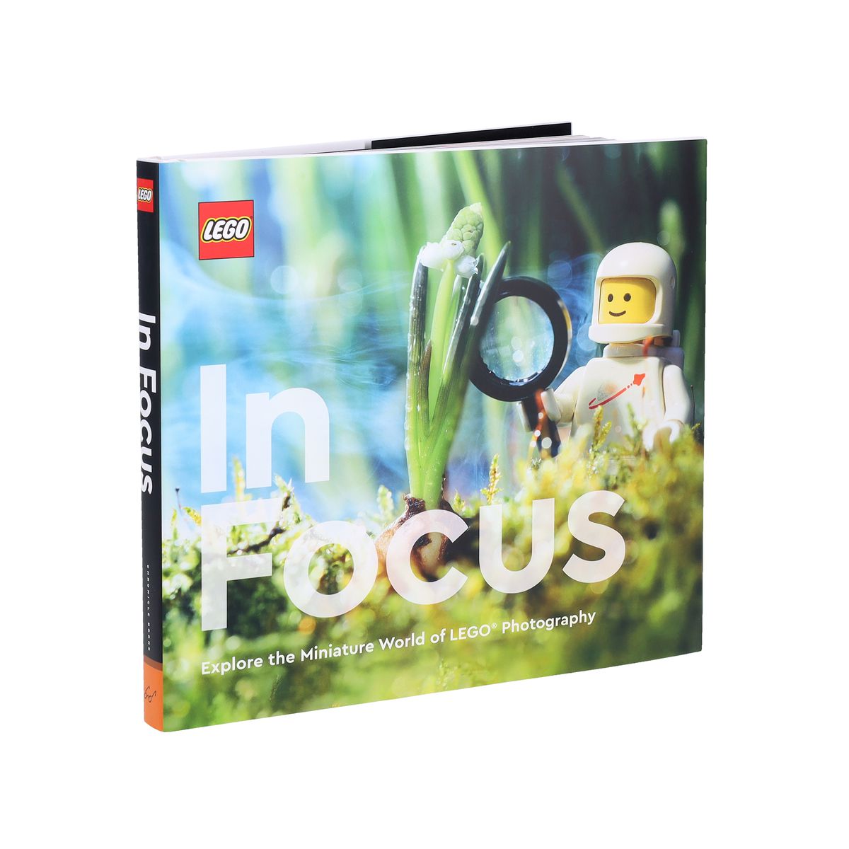 The cover to Lego in Focus, featuring a space helmet wearing man in white holding a magnifying glass looking at a small plant