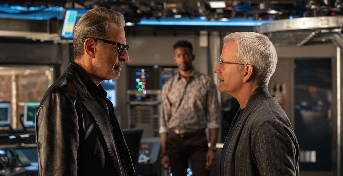 Dr. Ian Malcolm (Jeff Goldblum) and Lewis Dodgson (Campbell Scott) face off in the foreground of a tech-heavy room, gunslinger-style, while Ramsay Cole (Mamoudou Athie) looks on in the background in Jurassic World Dominion.