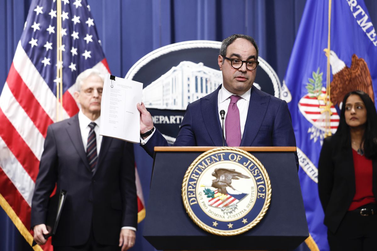 Jonathan Kanter holds up a document as he speaks alongside Attorney General Merrick Garland and Associate Attorney General Vanita Gupta at the Justice Department press podium with flags and a seal behind him. 