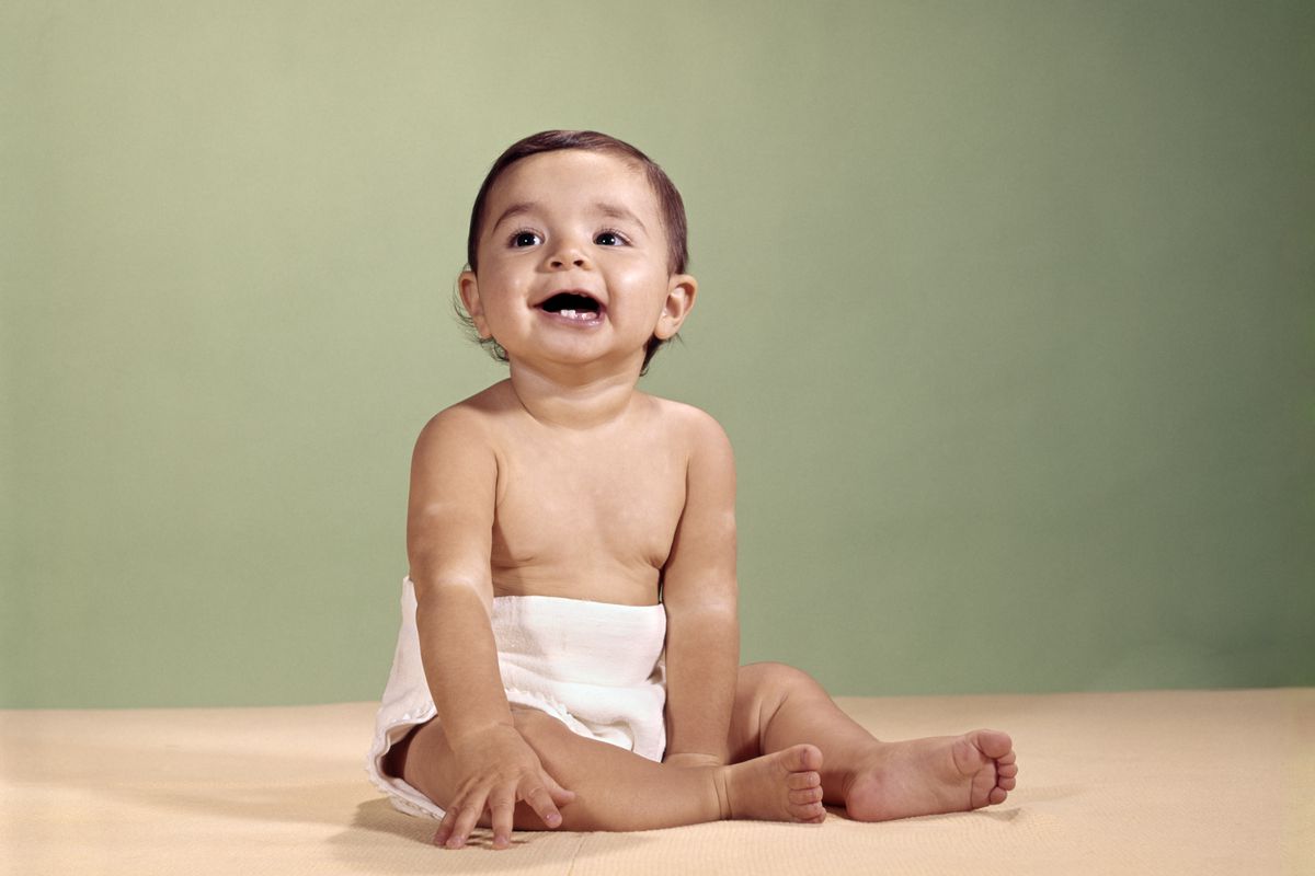 A cloth-diapered baby sitting on an empty floor and looking up smiling.
