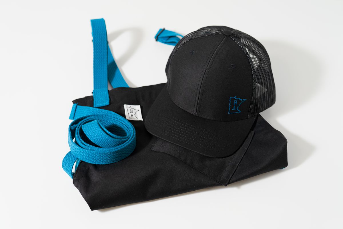 A folded black apron with blue strips and a black trucker hat with the logo stitched in blue thread