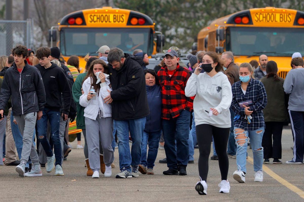 Students and family members walk on a black top as two school busses sit in the background.