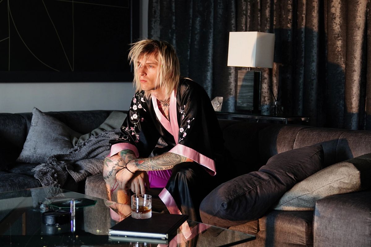 A photo of a young man covered in tattoos (Colson Baker/Machine Gun Kelly) in a black and pink bathrob sitting behind a glass living room table.
