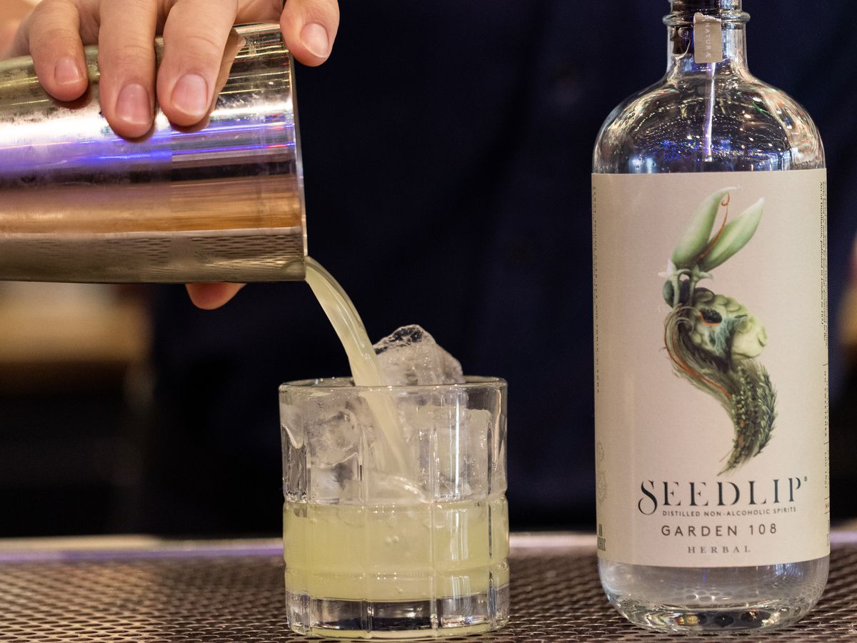 A bartender pours a pale green cocktail into a rocks glass, placed next to a bottle of Seedlip Garden 108.