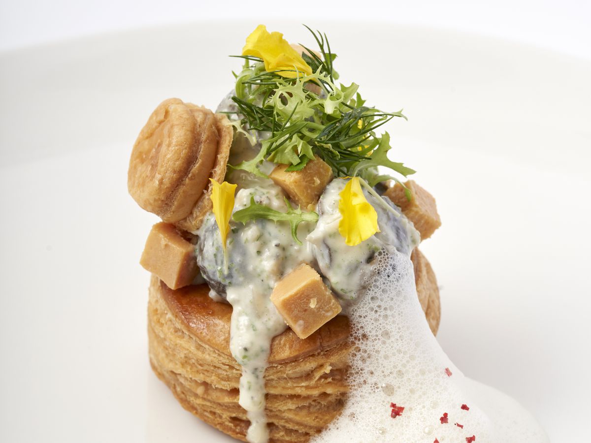 A sauce-covered oyster presented on top of a biscuit topped with greens, other fixings, and an explosion of foam.