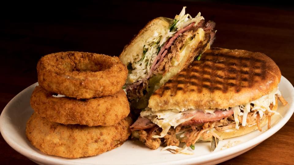 A pressed sandwich overflowing with meat and slaw, stacked next to a tower of onion rings. 