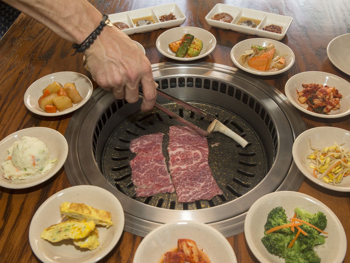 A person grilling meat on a tabletop grill that’s surrounded by banchan.