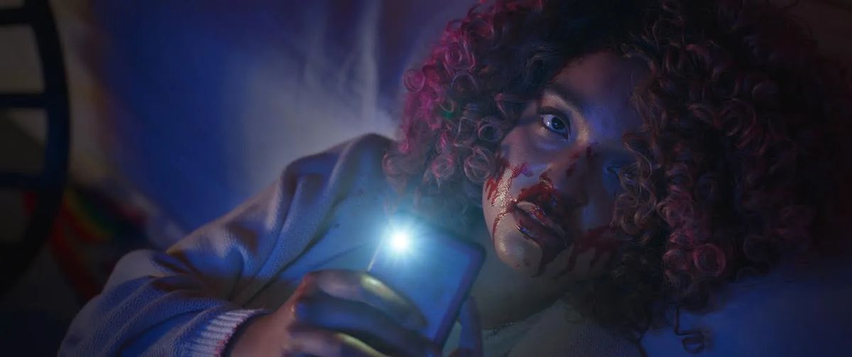 A woman with blood dripping down her nose and face with pink afro curls holds a cellphone in her hand with the camera light on.