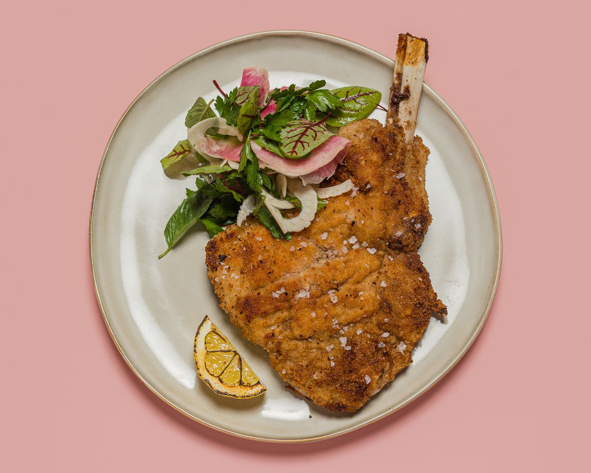 A bone-in chop of meat, fried, with lemon wedge at the side on a stone plate against a pink table at Mona Pasta Bar.