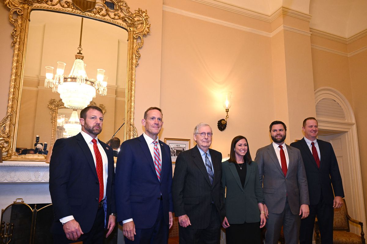 The senators stand in an ornate room with gilded chandeliers, sconces, and mirrors. They are in a line, all in black or navy suits, with the exception of Britt, in a forest green suit.