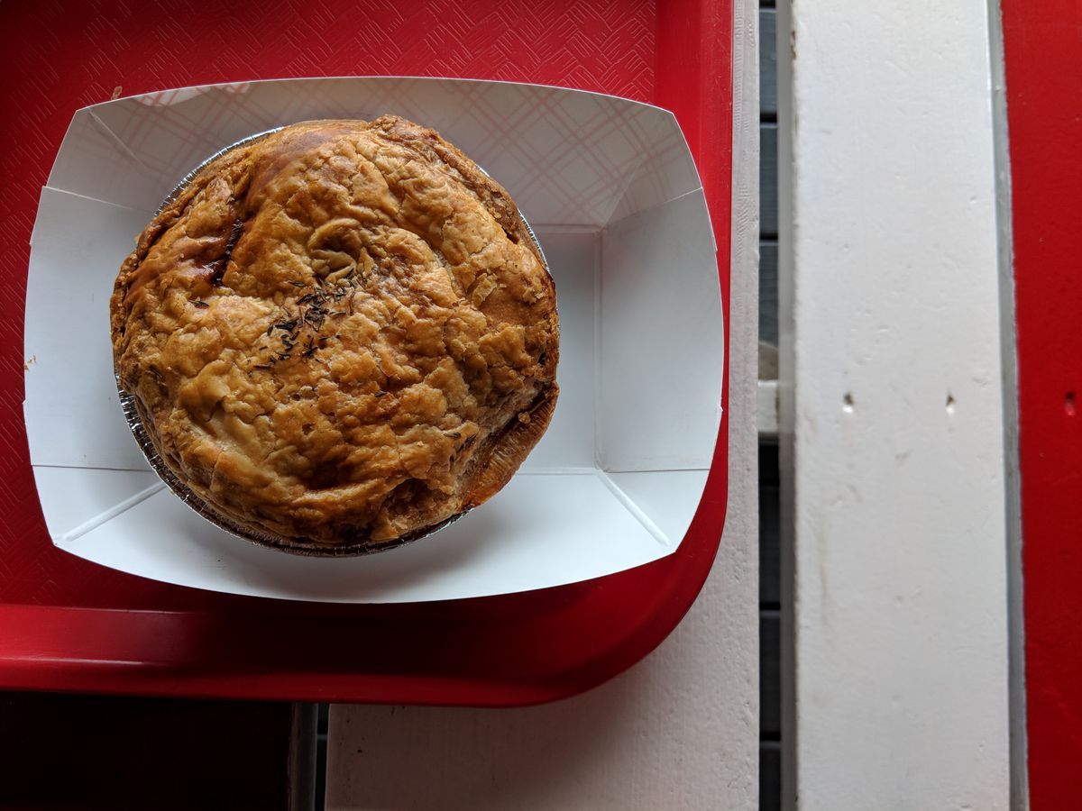 An Australian meat pie sits in a paper container on a red tray on a red and white picnic table