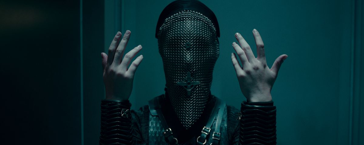 A warrior nun in a chainmail coif covering her face holds up her hands, palms inward.