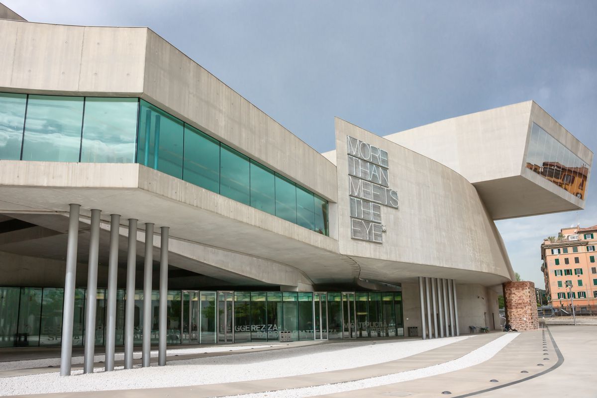 The exterior of the Maxxi National Museum in Italy.  The facade is gray and geometric. There is a neon sign on the side of the building that reads: More than meets the eye. 