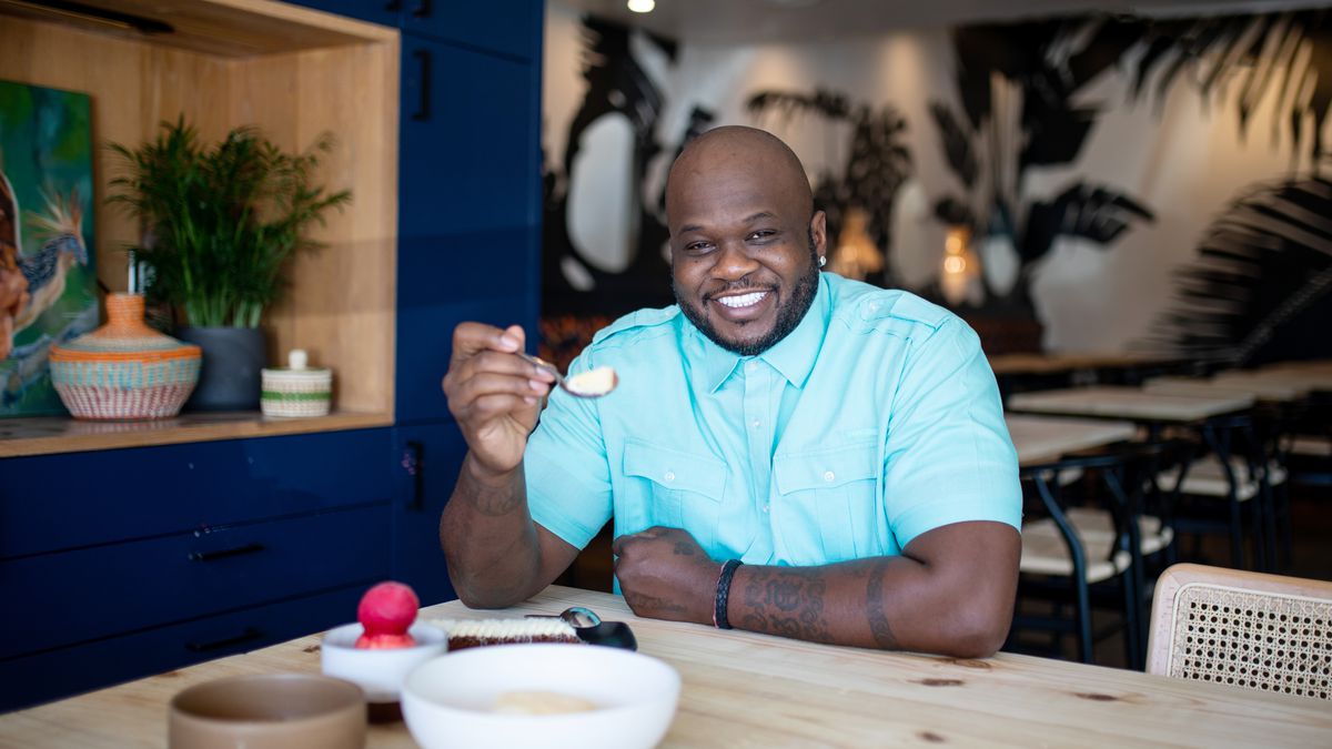 A man in a light teal button-down shirt seated at a table holding up a spoon of dessert and there’s a bowl of food on the table.