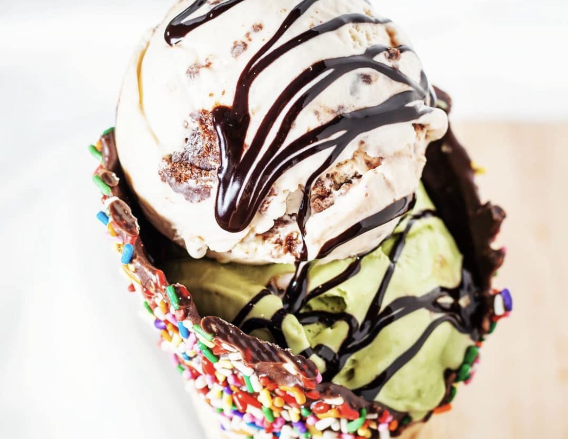 A close up of a double scoop of ice cream with chocolate sauce in a cone covered in rainbow sprinkles.
