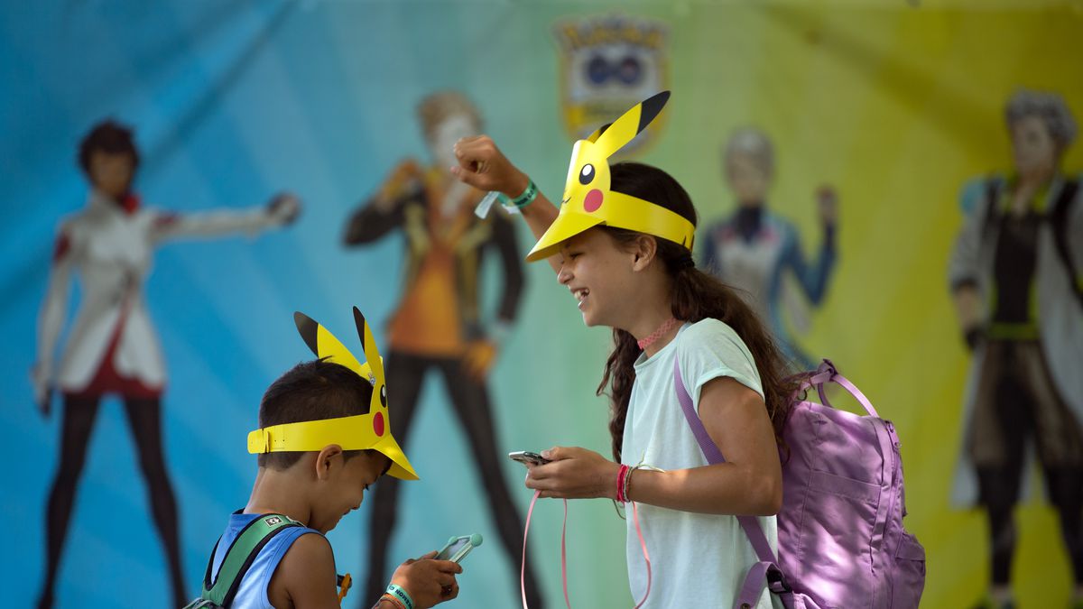 A photo of an 8-year-old and a 10-year-old brother and sister playing Pokémon Go on their phones. They are smiling and wearing special cardboard Pikachu visors.
