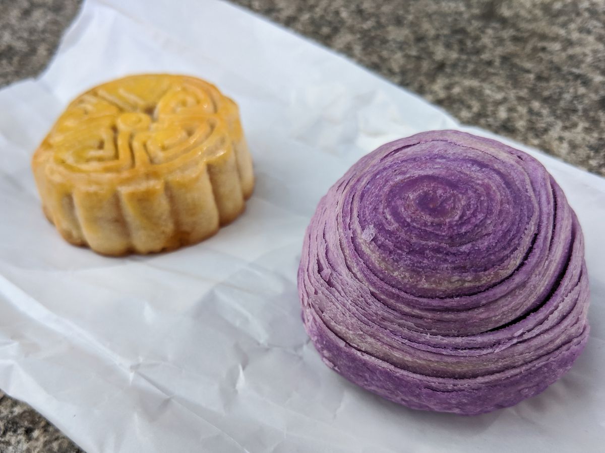 A swirled purple pastry made of taro and a small glossy mooncake sit on a white paper bag on a stone background