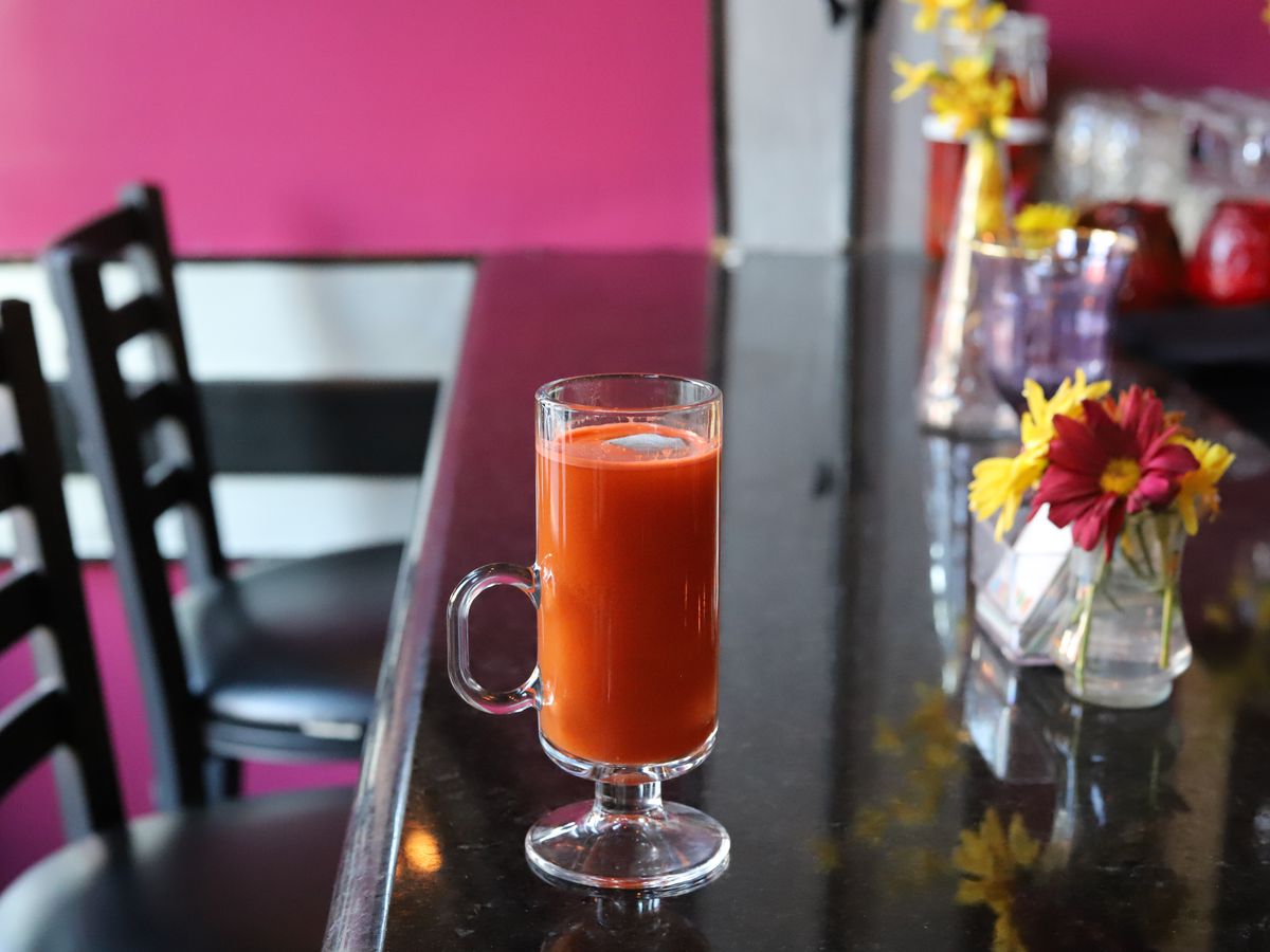 An orange-red hot cocktail made with butternut squash syrup and bourbon sits in a glass on a shiny black bar. There are flowers on the bar, and a pink wall is visible in the background.