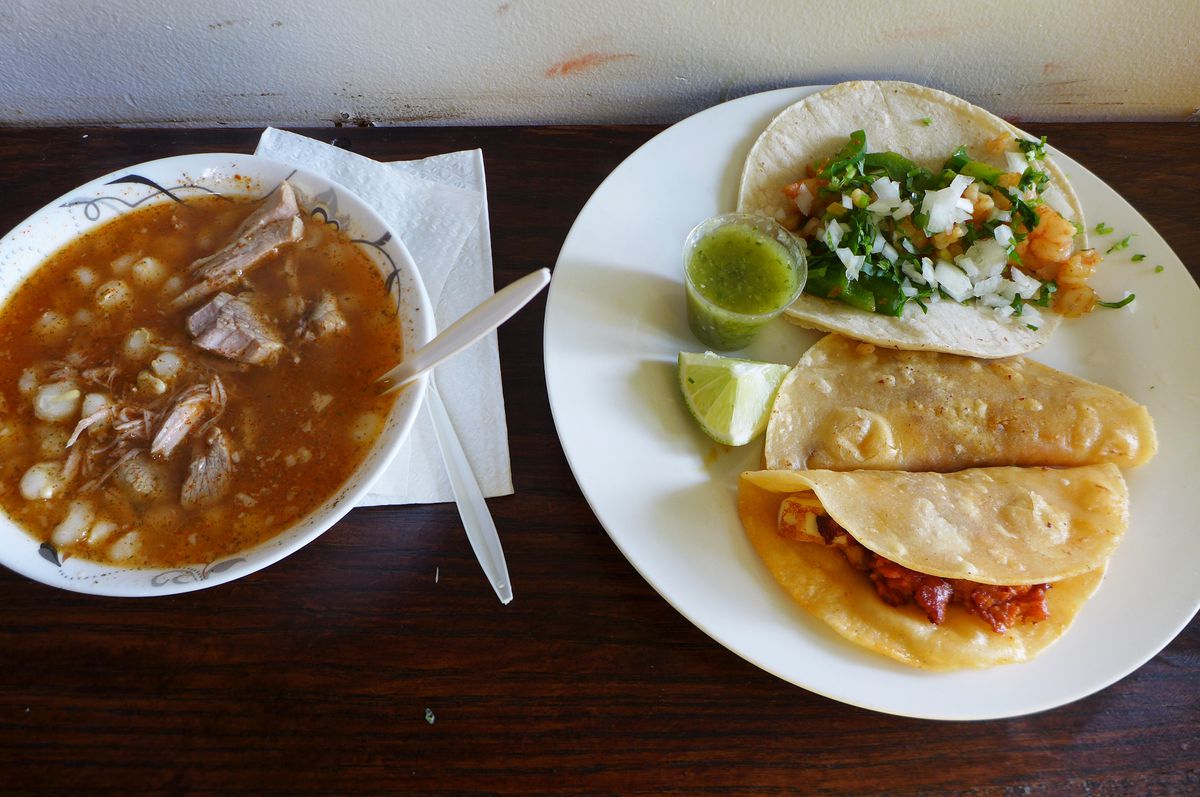 A white bowl of red pozole with onion and bobbing bits of meat, next to a plate full of tacos overflowing with shrimp.