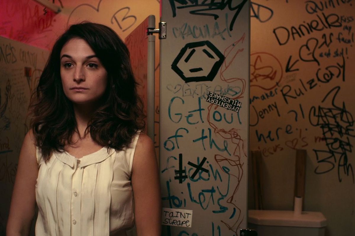 A young woman stands in a room, walls covered in graffiti.