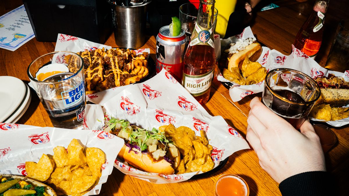 Hot dogs, burgers, beers, and colorful shots crowd a wooden table at a bar.