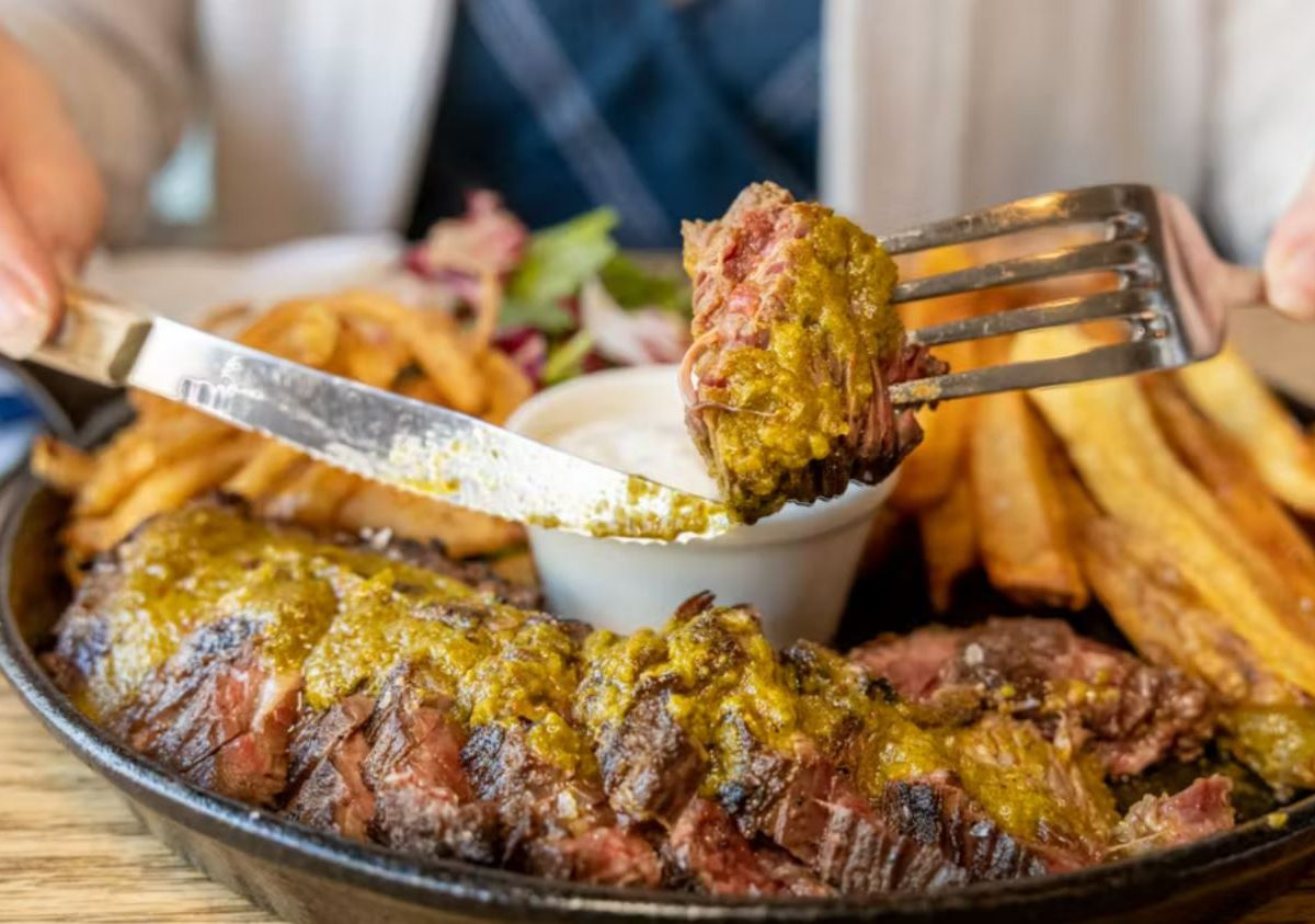 Two hands cut a giant steak smeared with yellowish sauce and surrounded by french fries.