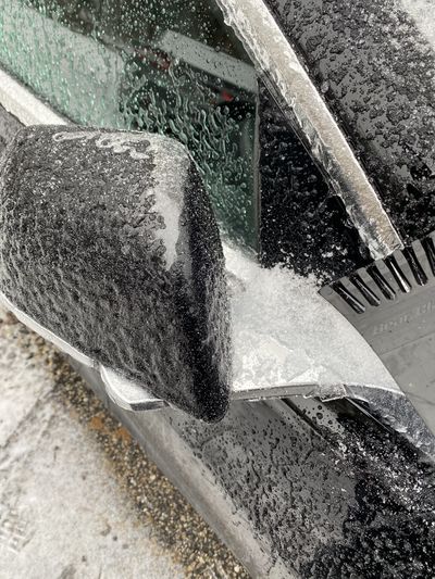 This is a closeup of the backside of a passenger side view mirror, which is folded and has ice around the joints. An ice scraper is being used to break it.